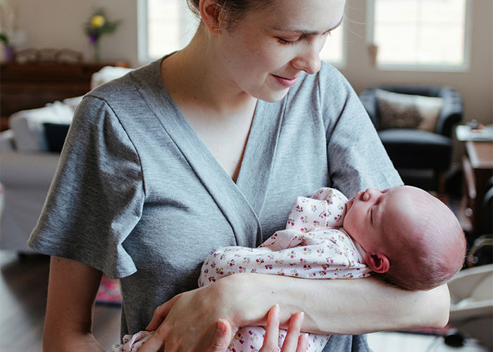 Woman in a gray shirt smiling while holding a newborn in a living room setting, highlighting baby names context.