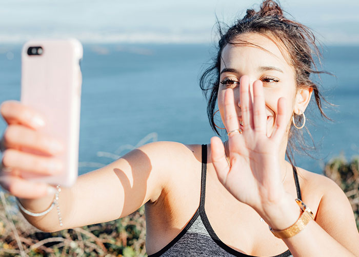 Woman taking selfie by the sea, smiling with raised hand; discussing baby names.