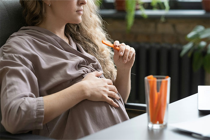 Pregnant woman in a relaxed pose holding a carrot stick, with a glass of carrot sticks on the table.