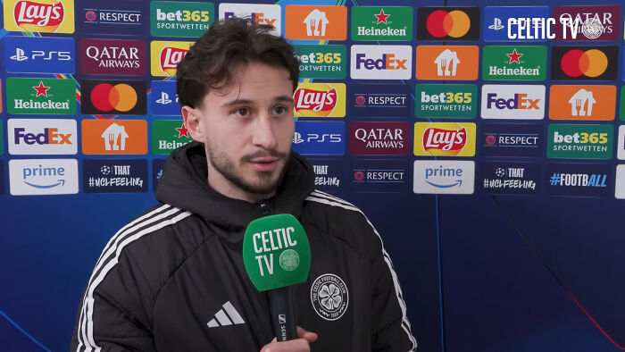 Celtic player giving a post-match interview with sponsor logos in the background, discussing the Champions League match against Bayern.