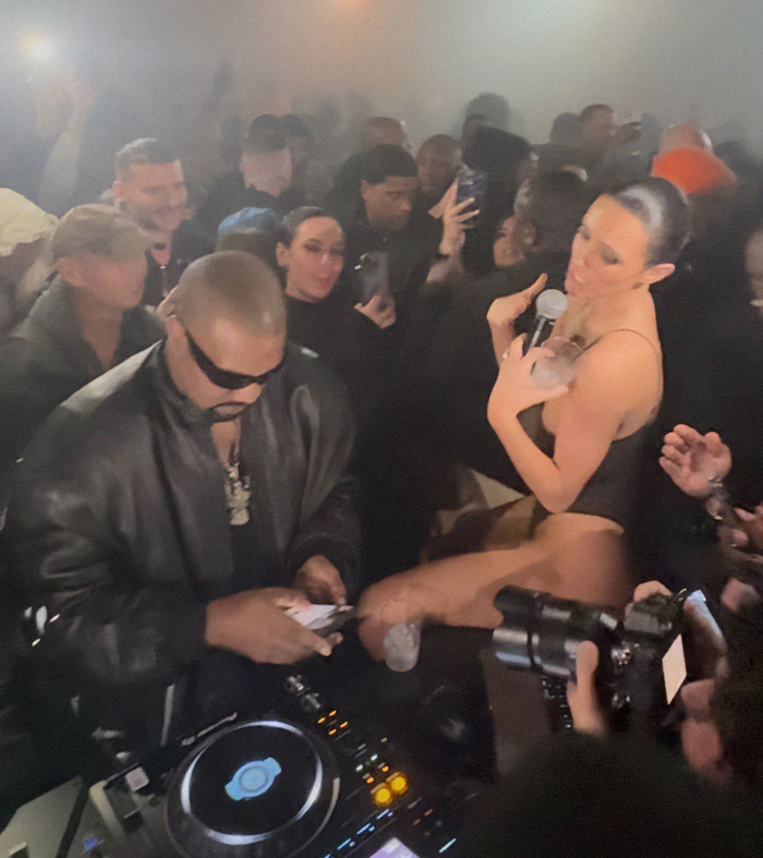 Bianca Censori at afterparty, holding a microphone, surrounded by a crowd in a dimly lit room.