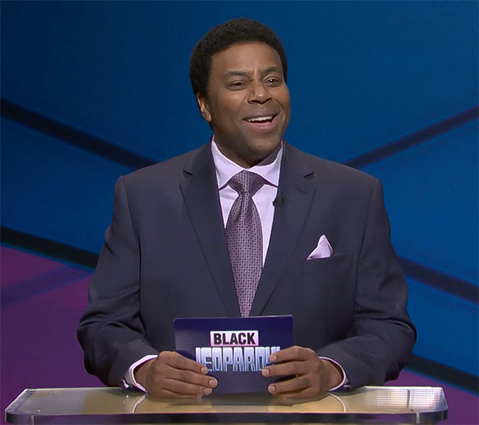 Host in a suit on a game show set, holding a card, related to SNL episode discussions.