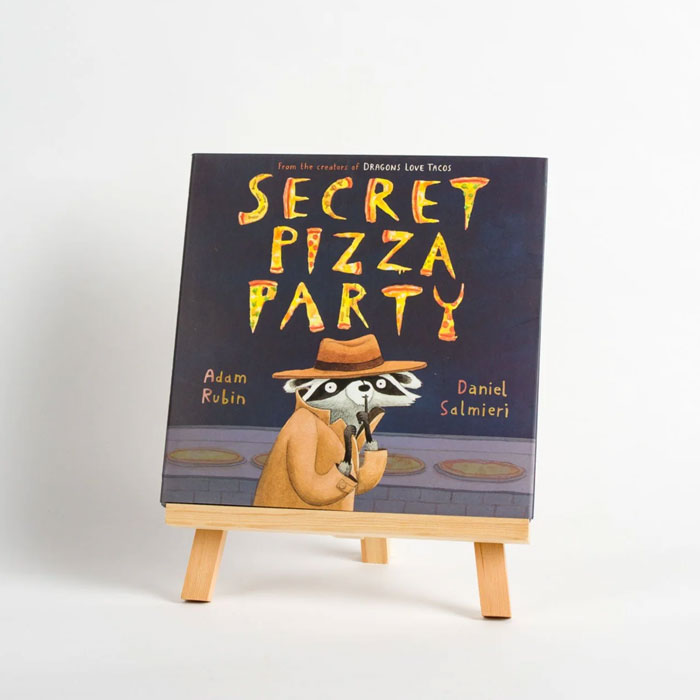 Secret Pizza Party" book displayed on an easel, featuring a raccoon wearing a coat and hat. Secret Pizza Party" book displayed on an easel, featuring a raccoon wearing a coat and hat.