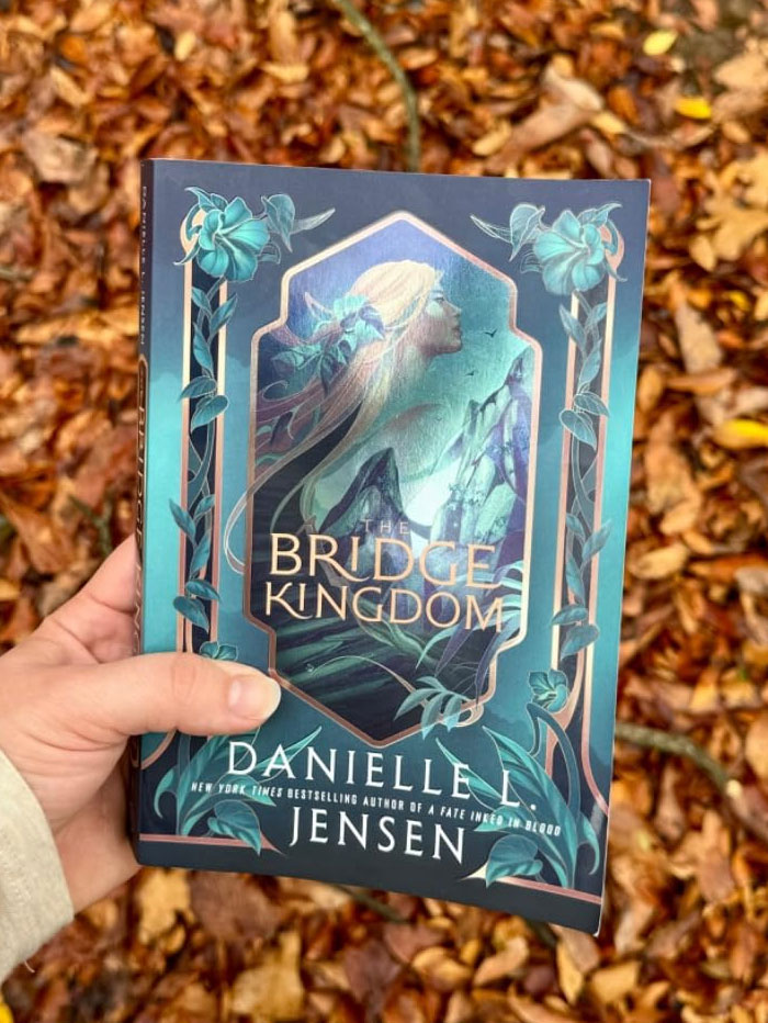 "The Bridge Kingdom by Danielle L. Jensen, held against a background of autumn leaves."