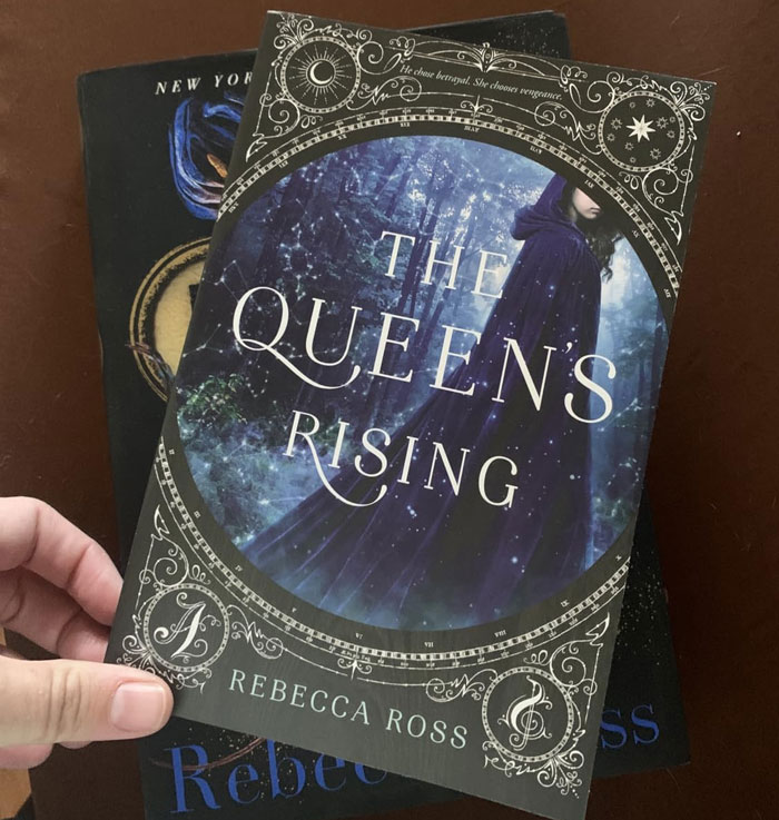 Book cover of "The Queen's Rising" by Rebecca Ross, held in hand.
