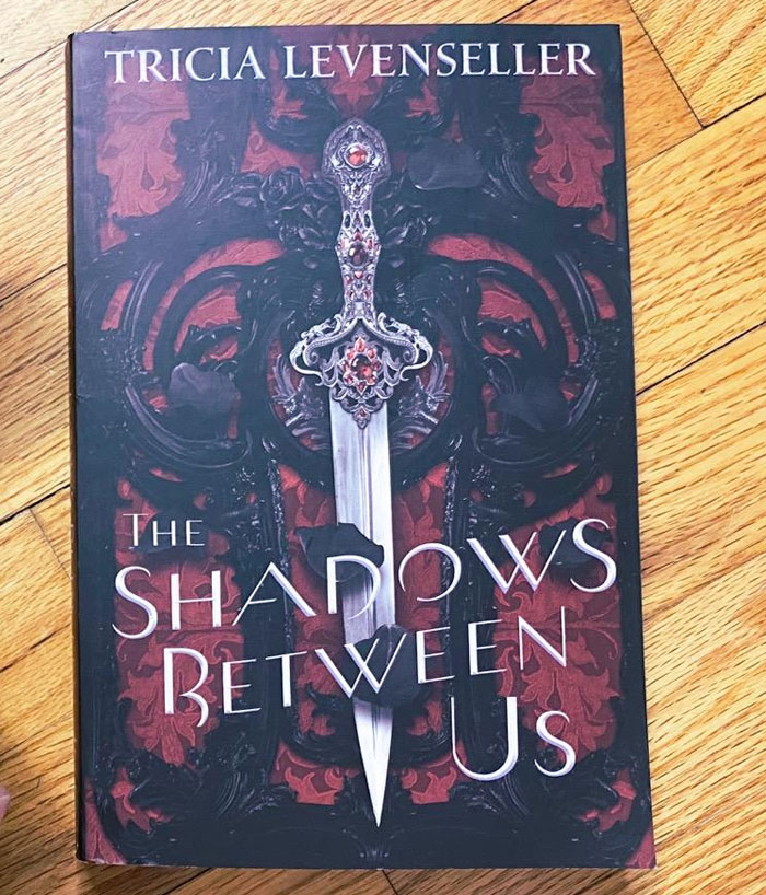 Cover of "The Shadows Between Us" by Tricia Levenseller on a wooden surface.