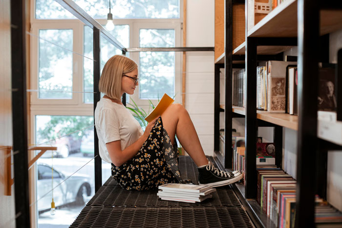 Woman reading a book in a library, surrounded by shelves, seeking support after novel planning setback.