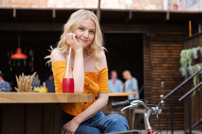 Blonde woman sitting at a cafe table with a red drink, appearing contemplative about novel planning frustrations.