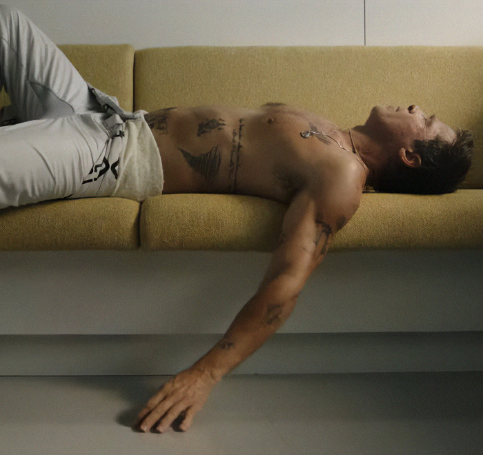 Shirtless man with tattoos relaxing on a yellow couch, showcasing body art.