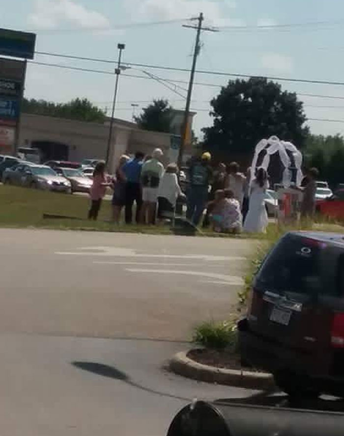 Group gathers for a budget wedding in a parking lot under makeshift arch on a sunny day.