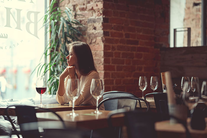 Woman sitting alone at a restaurant table with empty chairs, looking thoughtful, with a glass of red wine in front of her.