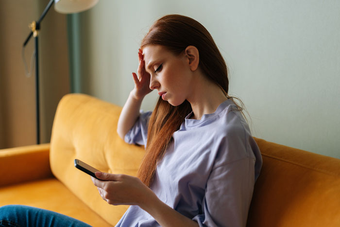 Woman upset after boyfriend cancels date, sitting on sofa and looking at phone.