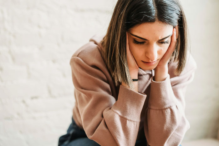 Woman looks upset after boyfriend cancels date, sitting in a beige sweater, head in hands.