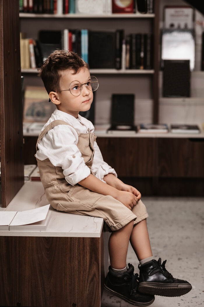Young boy wearing glasses and beige overalls sitting thoughtfully in a cozy office, capturing an insane workplace moment.