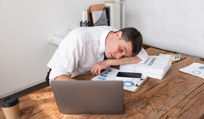 Man overwhelmed at wooden desk with laptop, financial reports, and phone, depicting insane workplace moments.