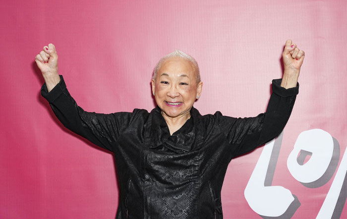 Lori Tan Chinn in black jacket smiling with hands raised, against a pink background, cast member of Netflix series "A Man On The Inside".