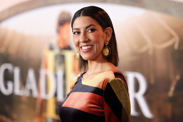 Stephanie Beatriz, a cast member of Netflix's "A Man On The Inside" at a public event, wearing a multicolored dress and gold earrings.