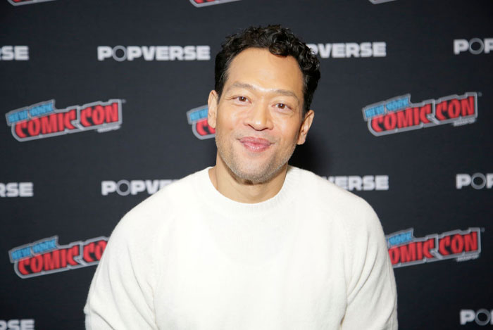 Eugene Cordero from A Man On The Inside cast at a Comic Con event, wearing a white shirt, smiling in front of Popverse backdrop.