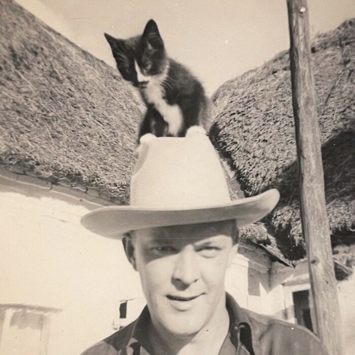 Black and white photo of a man wearing a hat with a kitten perched on top, capturing a vintage-cat-photo moment.