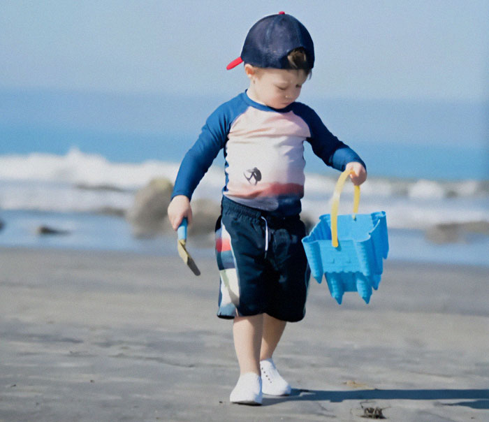 A young boy on the beach with a blue bucket and spade, wearing a cap.