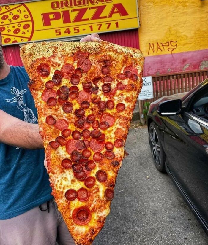 Huge pepperoni pizza slice held in front of a yellow pizzeria sign, showcasing common USA things.