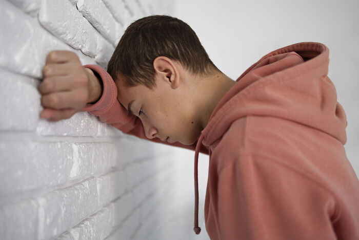 Young person in a pink hoodie, resting head against a white brick wall, expressing emotion related to feeling loved.
