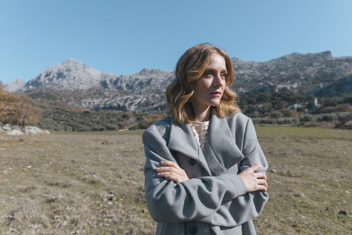 A woman in a gray coat standing in a mountain landscape, contemplating the harsh realities about life.