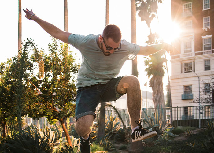 Hombre saltando al aire en camiseta y pantalones cortos en parque urbano al atardecer, mostrando rasgos poco atractivos.