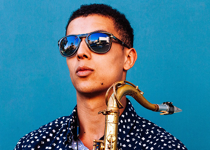 Joven con rasgos poco atractivos tocando saxofón, luciendo gafas de sol y camisa estampada ante fondo azul.
