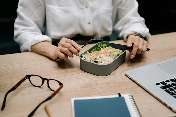 Person in white shirt eating lunch at a desk with a laptop, notebook, and glasses nearby. Person in white shirt eating lunch at a desk with a laptop, notebook, and glasses nearby.