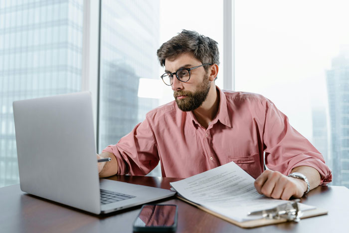 Coworker in pink shirt at desk, working on laptop and holding document, illustrating office setting.