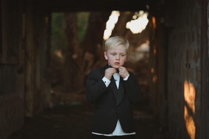 A young boy in a suit stands in a dimly lit passage, adjusting his bow tie, with a serious expression.