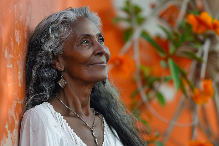 Elderly woman smiling beside an orange wall adorned with vibrant flowers.