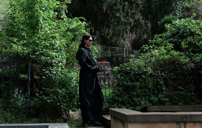 Person in black coat holding a flower in a lush, green cemetery.
