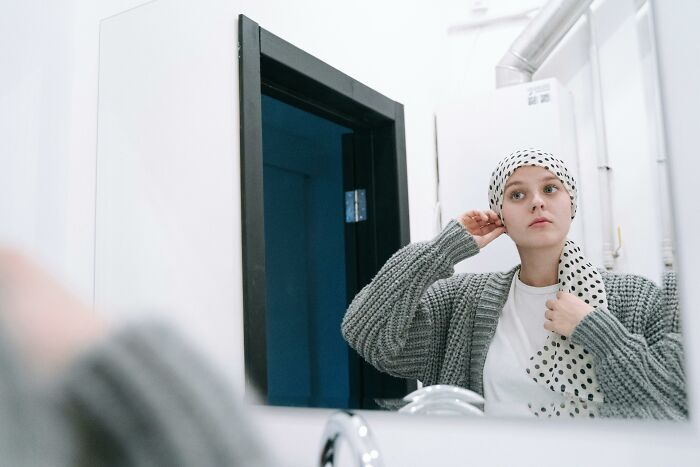 Person in a gray sweater adjusting a polka dot headscarf, standing in front of a mirror, possibly feeling reflective or anxious.