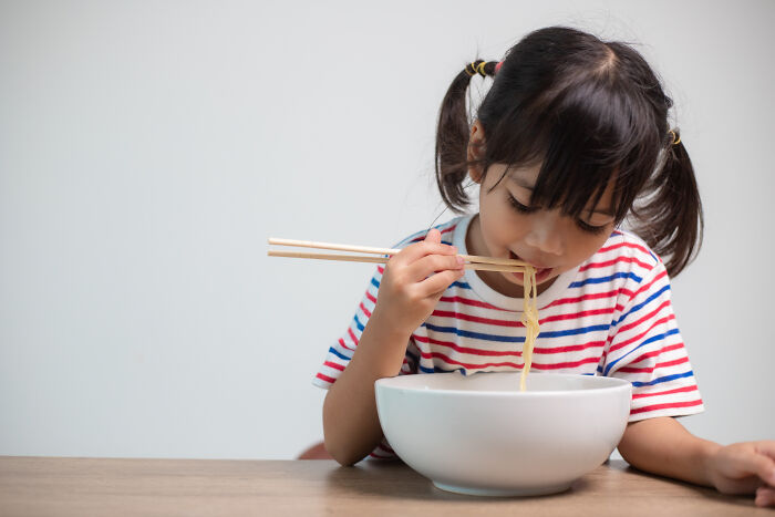 Child in a striped shirt eating noodles with chopsticks, showcasing the innocence often preceding creepy things kids might say.