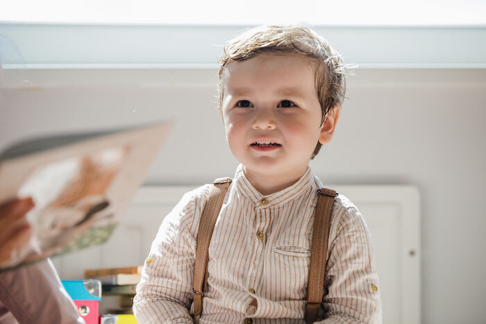 A young child in a striped shirt looks thoughtful, sitting indoors with sunlight streaming in.