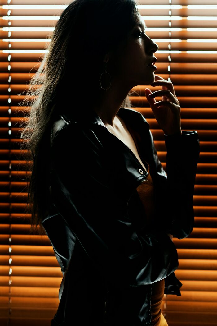 Silhouette of a woman with a prominent nose, standing against window blinds, in a thoughtful pose.