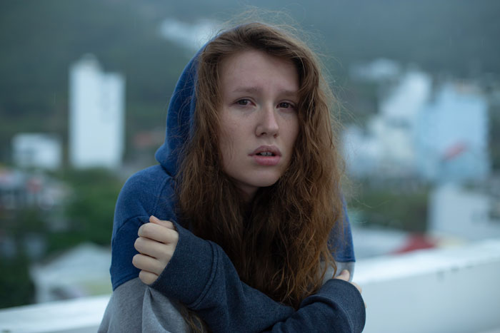 Teen with long hair and blue hoodie looks concerned, sitting outdoors with a blurred urban background. Teen with long hair and blue hoodie looks concerned, sitting outdoors with a blurred urban background.