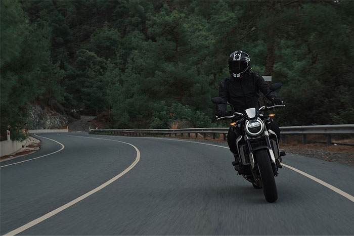 Motorcyclist wearing black gear on a winding road through dense forest, evoking a sense of mystery and dark stories.