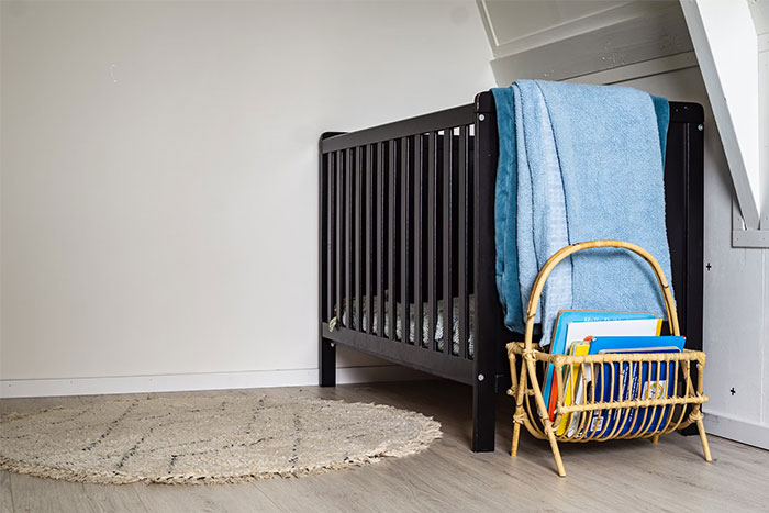 Empty crib next to a wall with blue blankets and a basket of books; evokes a haunting atmosphere.