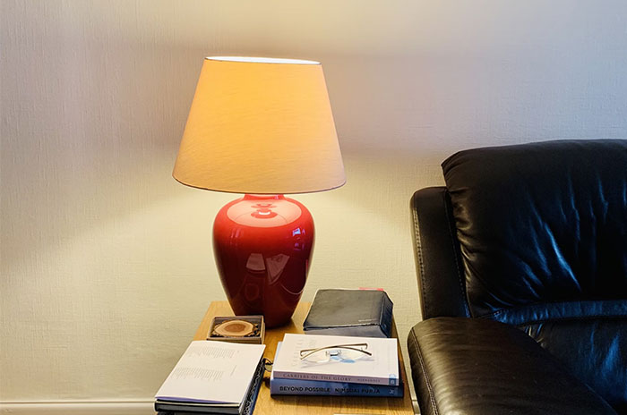 Red lamp on wooden table beside a leather couch, surrounded by books, capturing a cozy yet haunting atmosphere.