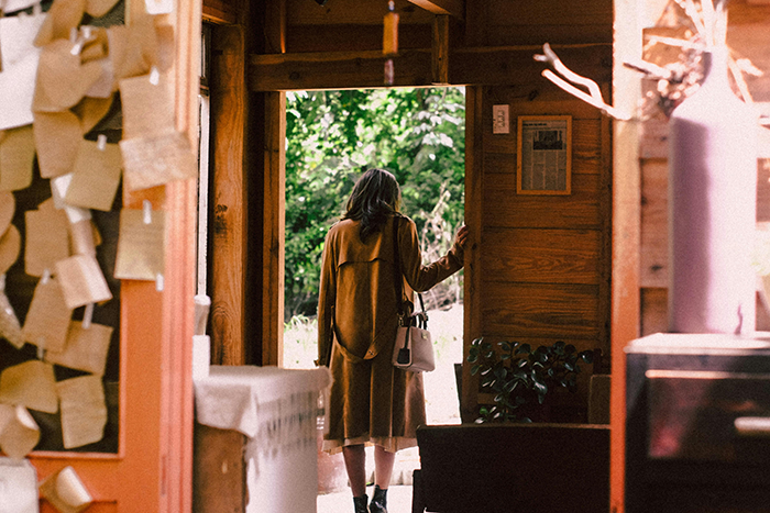 Person leaving a wooden cabin on a sunny day, holding a handbag, with trees visible outside. Person leaving a wooden cabin on a sunny day, holding a handbag, with trees visible outside.