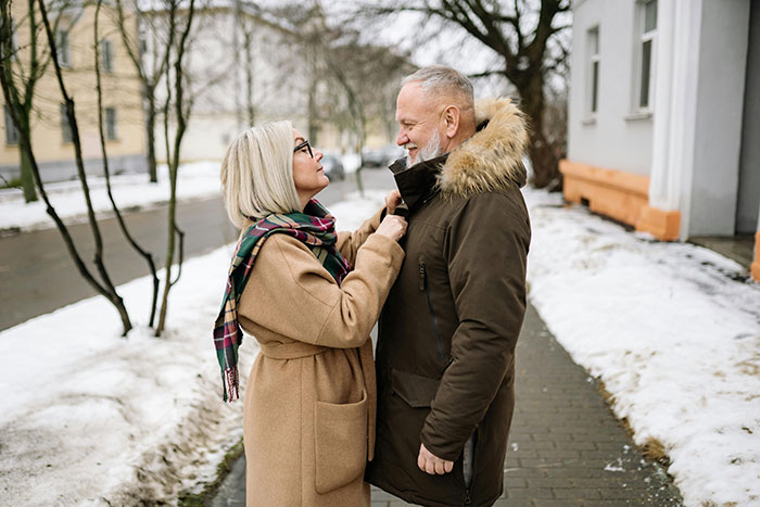 A woman adjusts a man's coat collar on a snowy sidewalk.