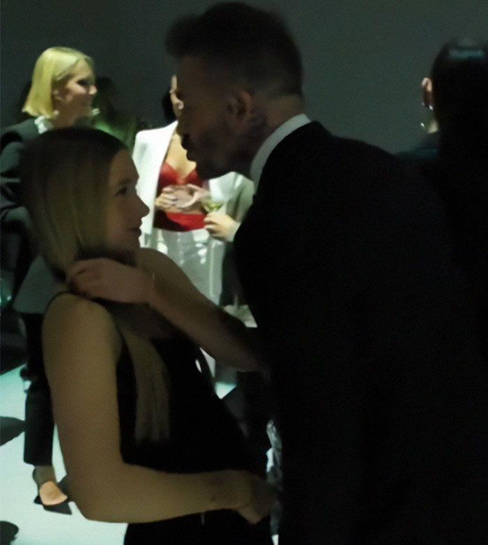 Man in a suit leaning towards a young girl in a dimly lit gathering, surrounded by people chatting. Man in a suit leaning towards a young girl in a dimly lit gathering, surrounded by people chatting.