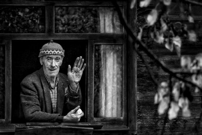 Elderly man in a hat waving from a window, captured in rural Turkey in striking black-and-white photography.