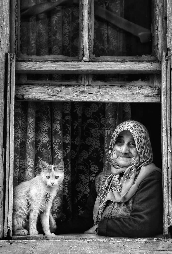 Elderly woman with a headscarf and a cat at a rustic window, showcasing rural Turkey in black-and-white photography.