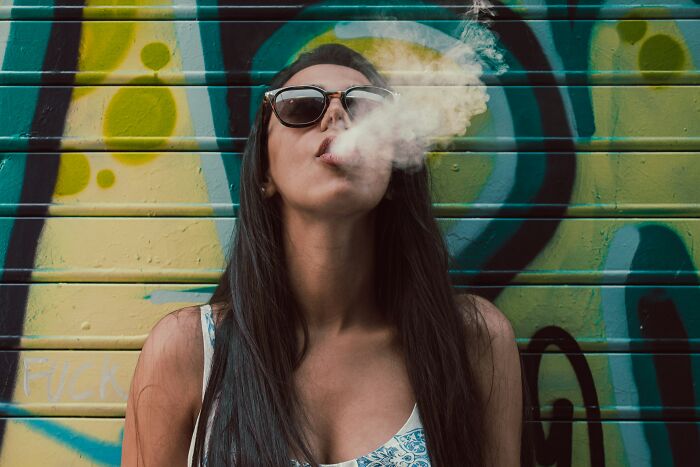 Woman exhaling smoke against a graffiti wall, wearing sunglasses; unforgettable moment captured.