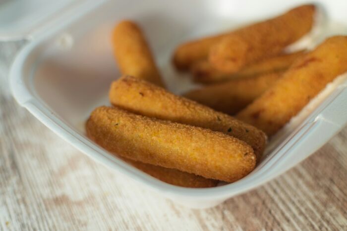 Golden breaded mozzarella sticks in a plastic container on a wooden table, indicating a deeply weird snack choice.