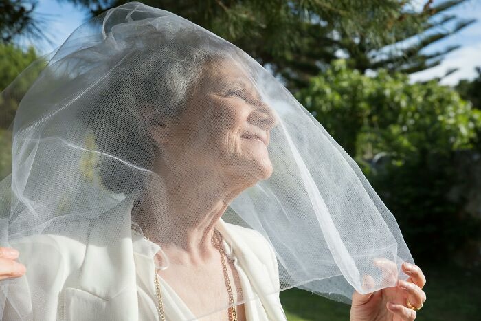 Elderly woman in wedding attire smiling under a veil, outside in natural light.